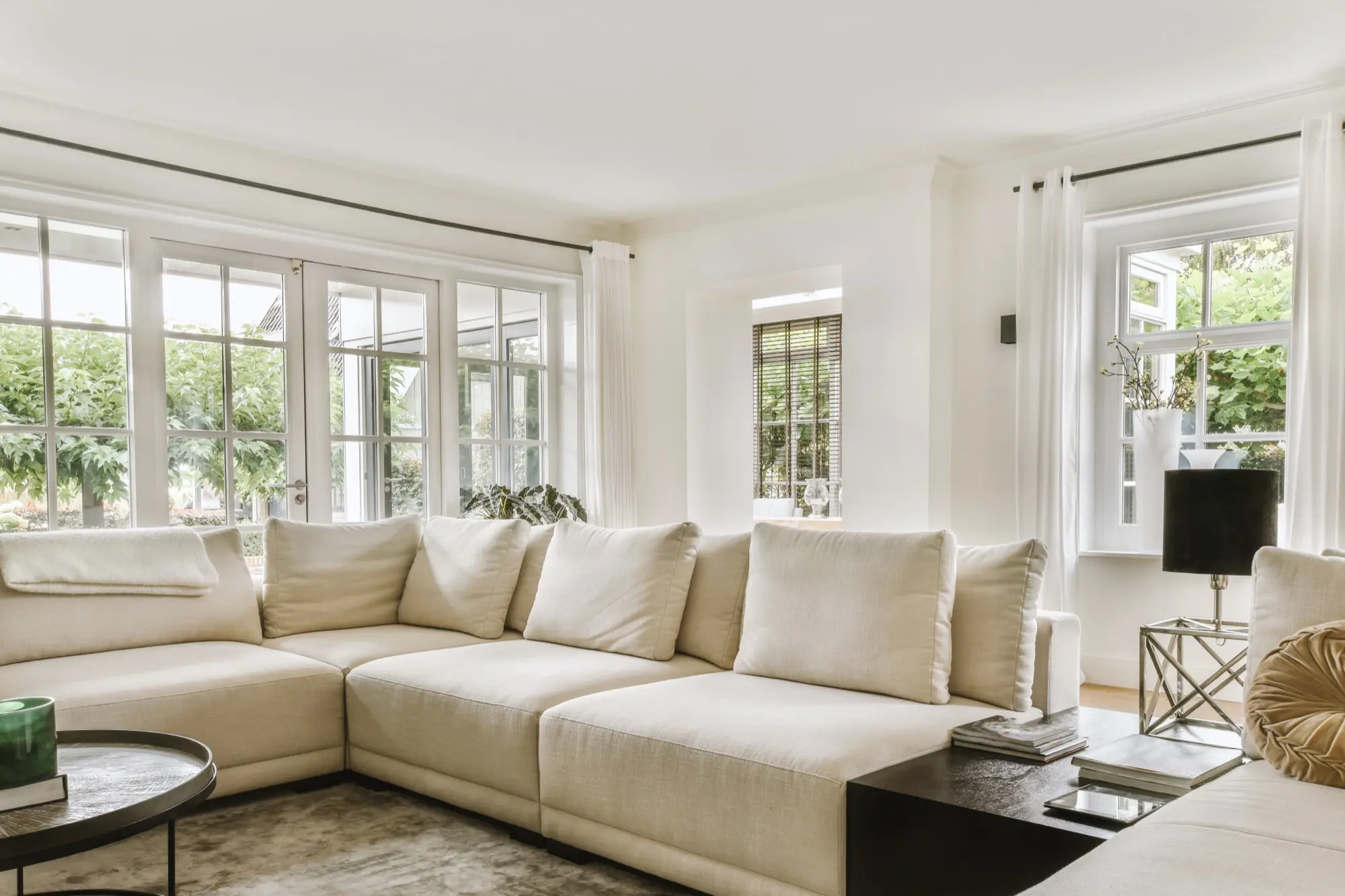 Helles Wohnzimmer mit beiger Couch, großen Fenstern und Pflanzen im Hintergrund.