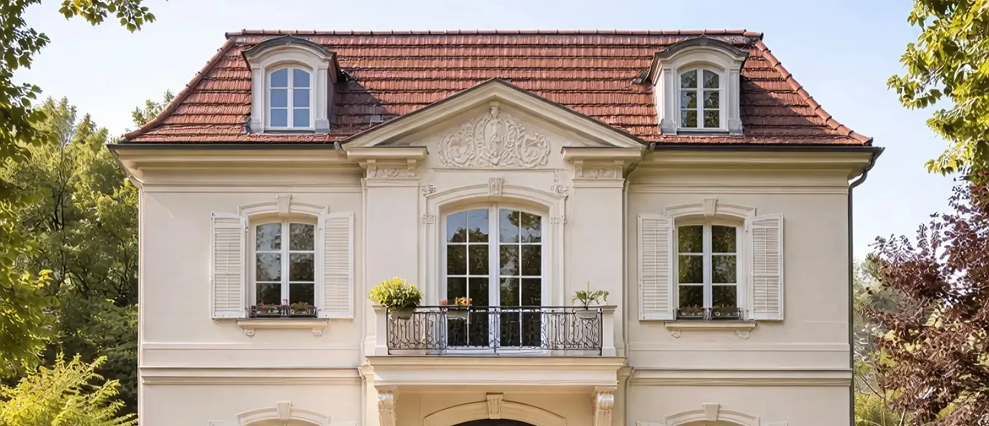Helles Gebäude mit rotem Ziegeldach, weißen Fenstern und Balkon, umgeben von Bäumen.